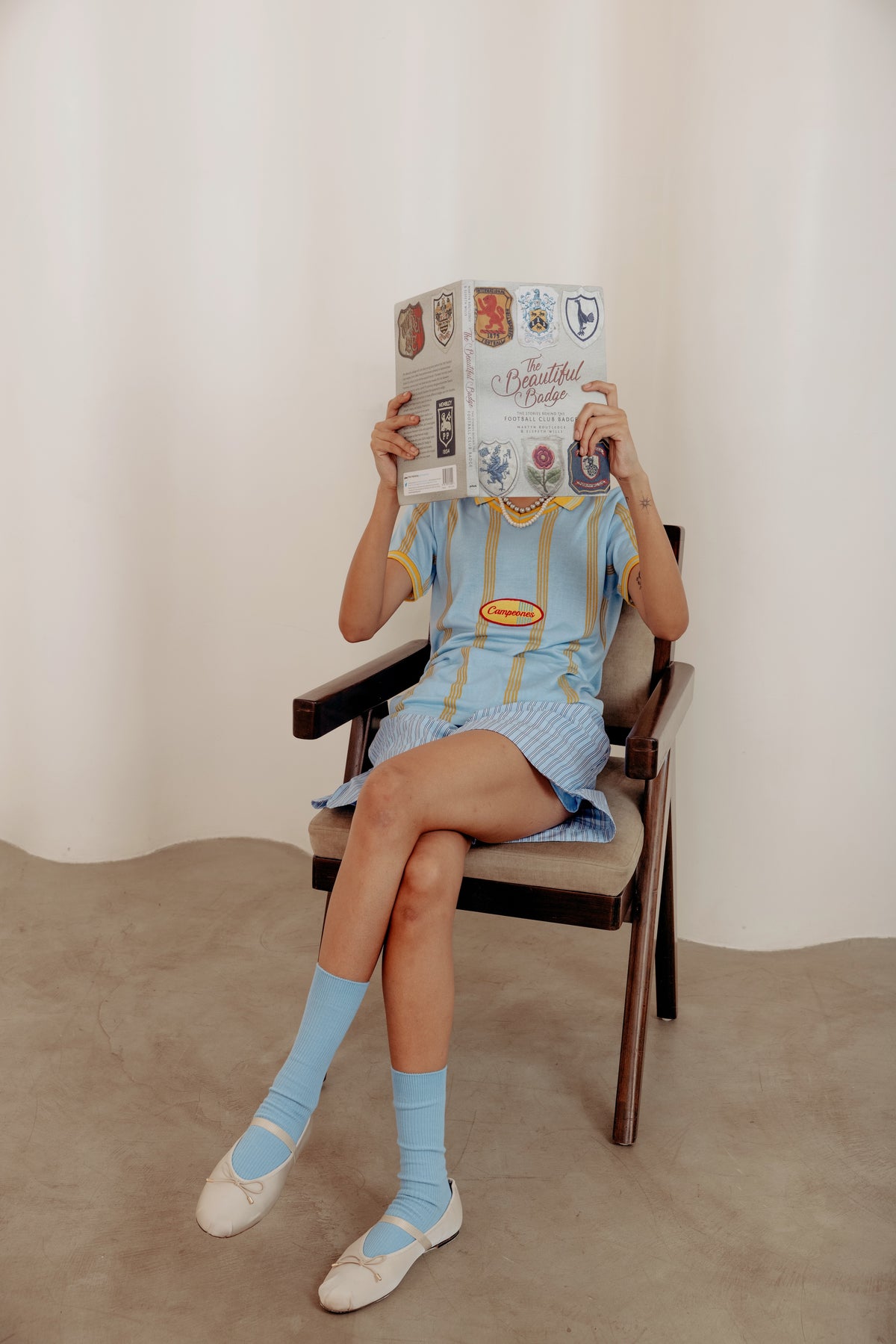Person sitting on a chair holding a book over their face, wearing a blue dress and socks.