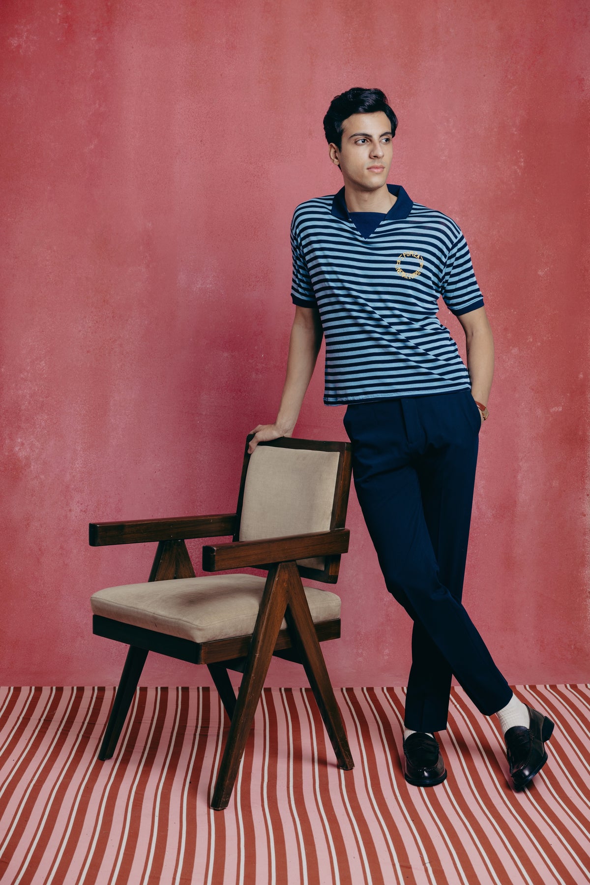 Man in a striped shirt and dark pants standing next to a chair against a pink wall.